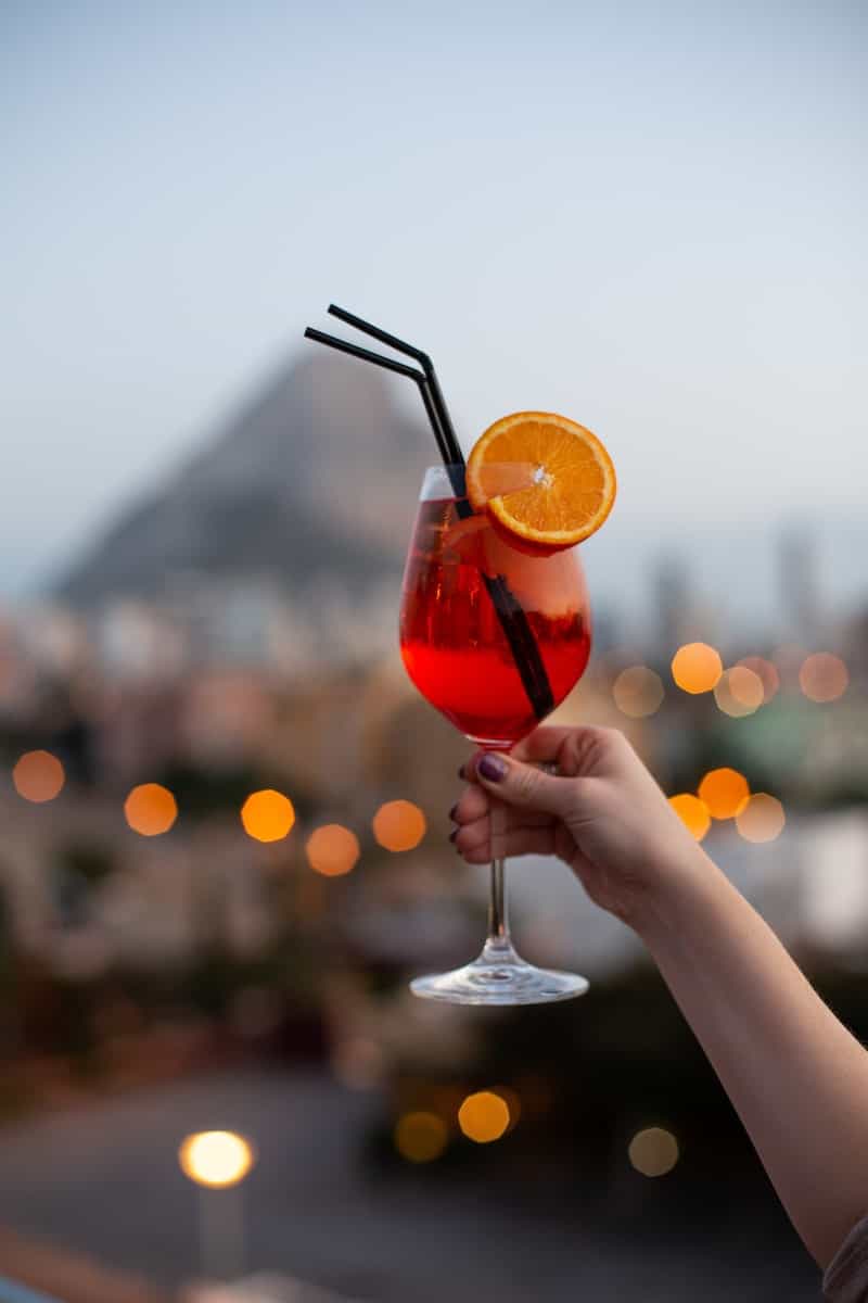 person holding clear wine glass with red aperol spritz and orange slice