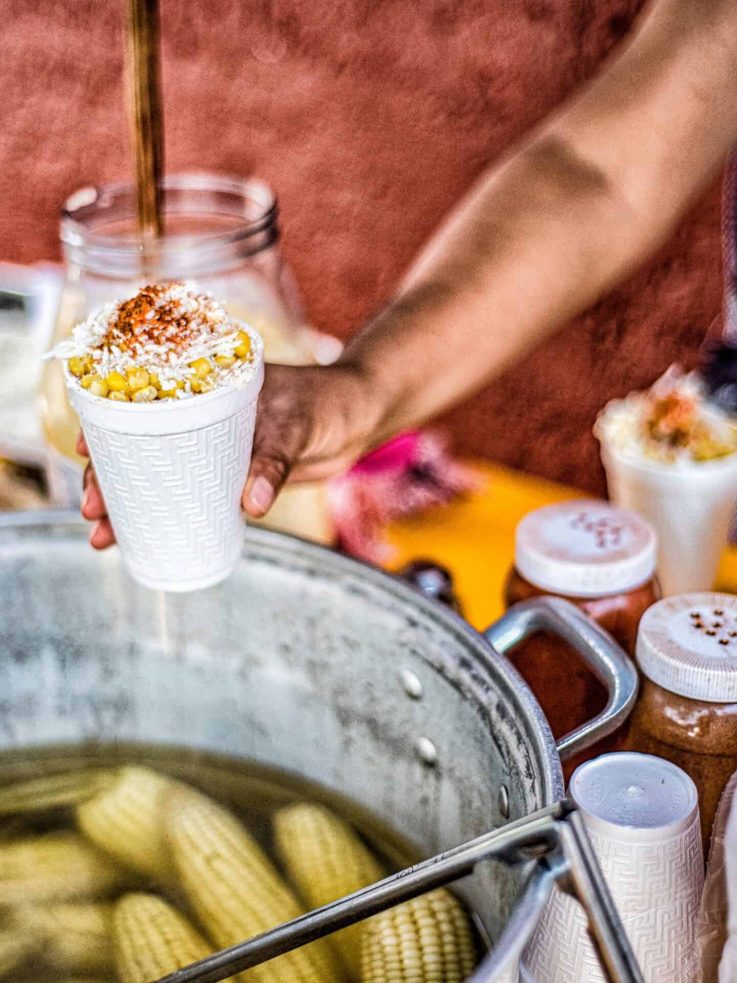 street vendor offering white cup of esquites over a boiling put of corn on the cob