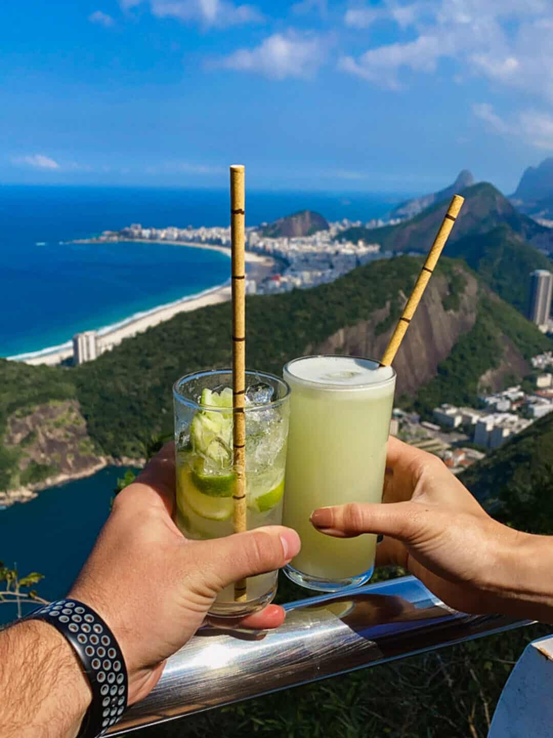 persons holding two yellow and clear alcoholic drinks on sugar loaf mountain overlooking city of Rio de Janeiro