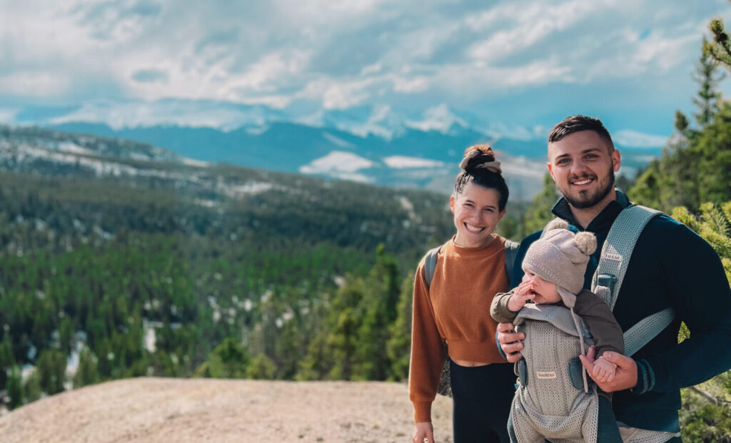 mother and father with baby strapped to chest on a hike in the rocky mountains under clear blue sky