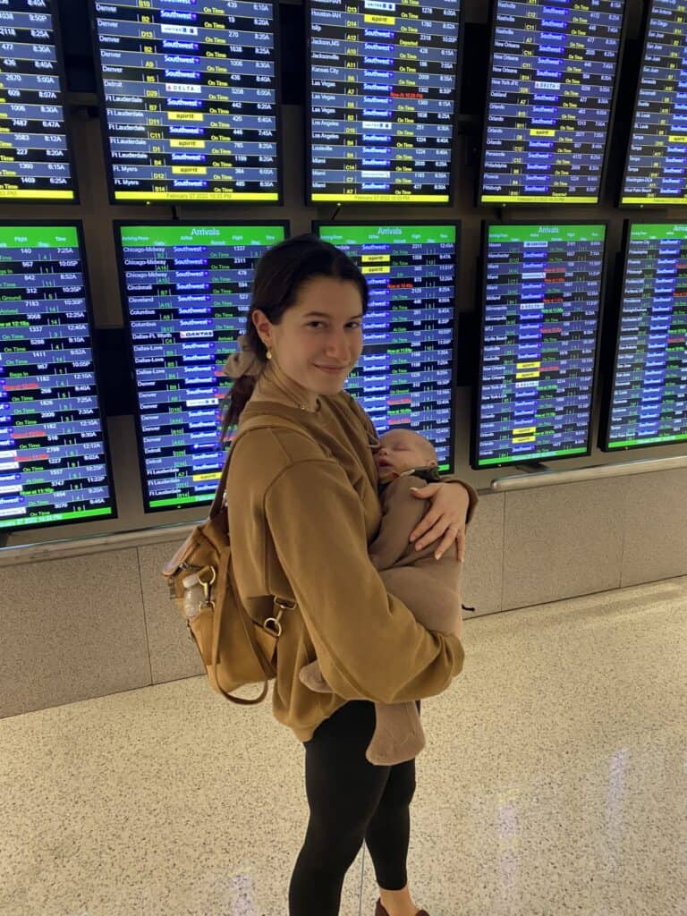mother holding sleeping baby in airport in front of departure screens
