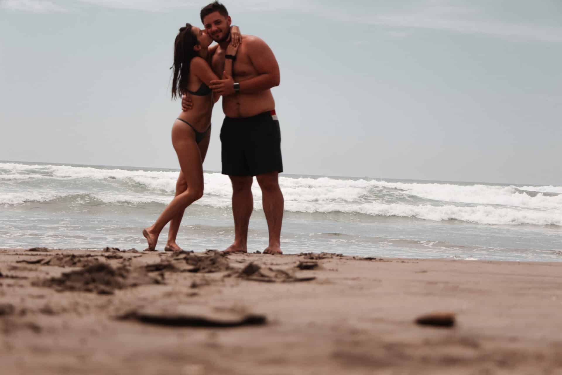 happy couple embracing and kissing on cheek under overcast beach sky