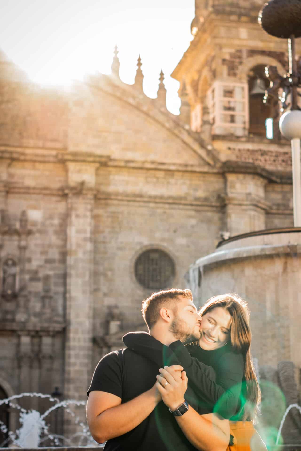 couple in black embracing and kissing underneath soft sunlight of colonial cathedral