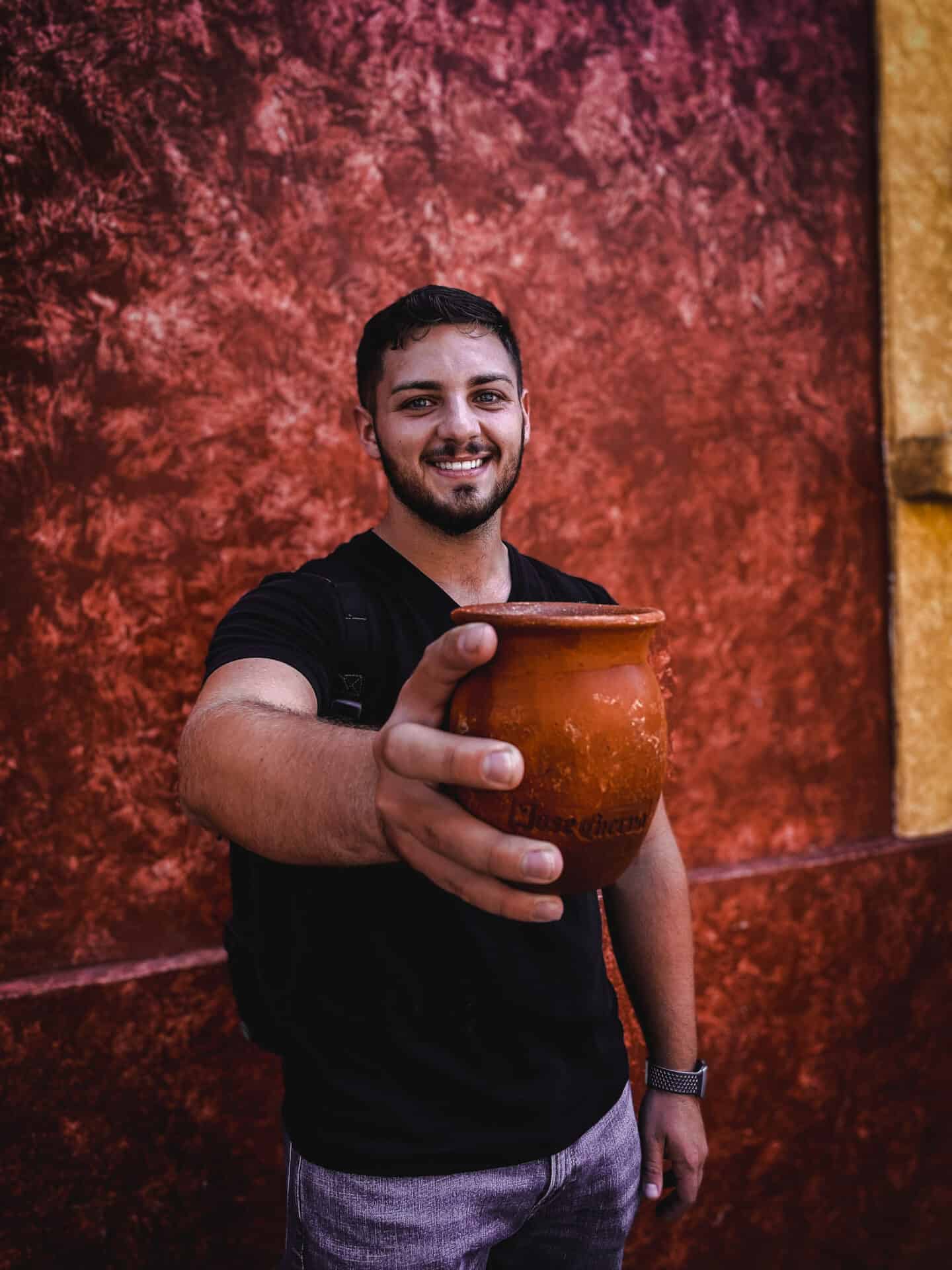 man wearing black shirt in front of red concrete wall holding up a clay canteen of alcohol