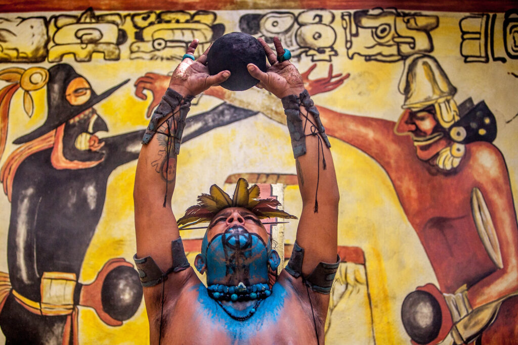 Mayan Man in makeup holding ball in front of mural