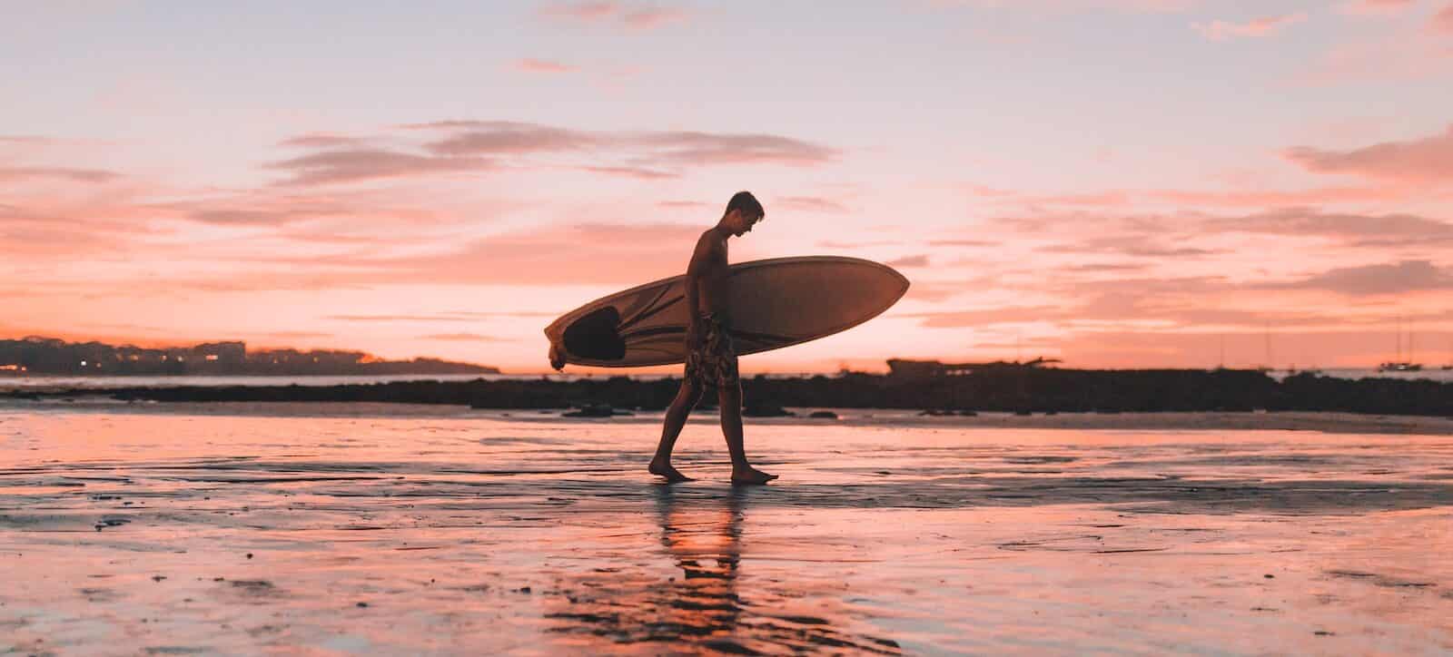 man holding surfboard walking near seashore under pink sky