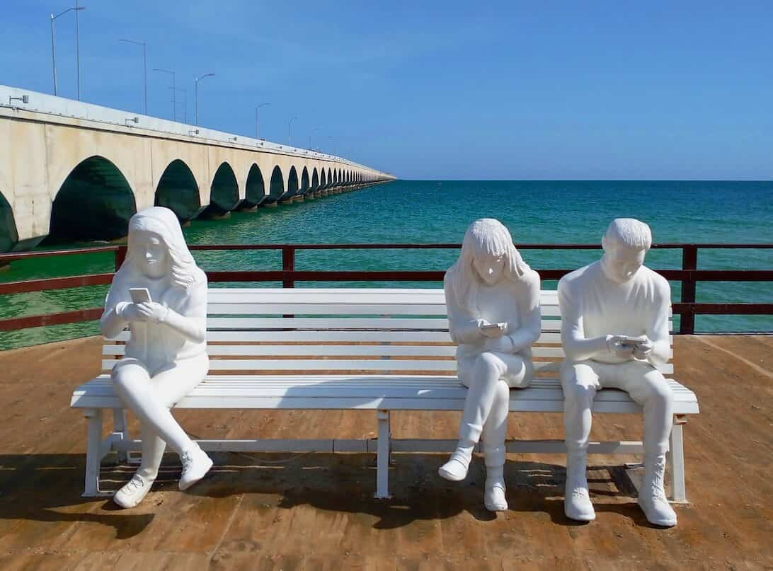 a pier with white bench with art statue people on their phones