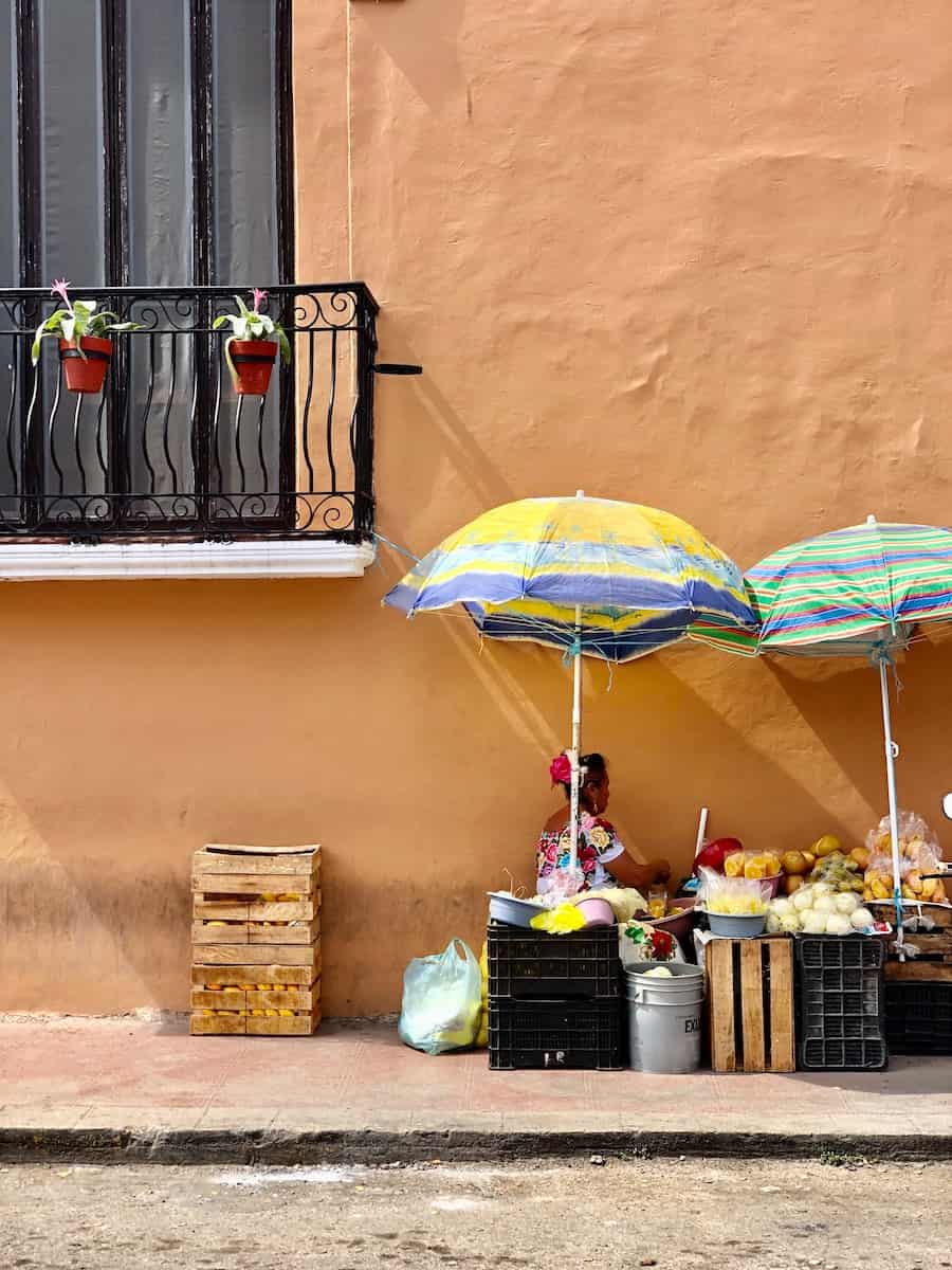 mexican woman sitting under umbrella next to peach colored building