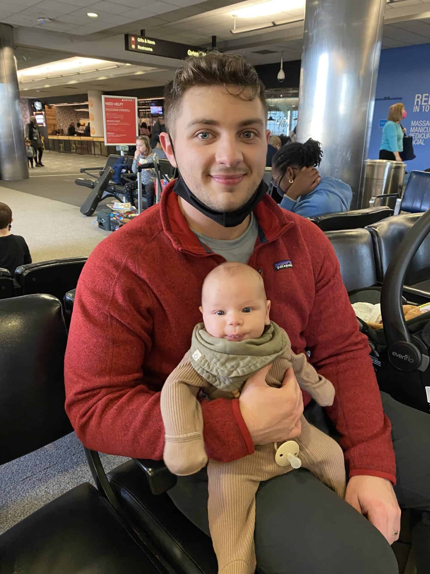 father in red sweatshirt holding two month old baby on his lap in an airport