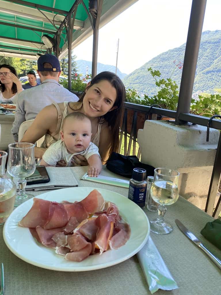six month old baby sitting on mothers lap at restaurant with large plate of prosciutto on the table