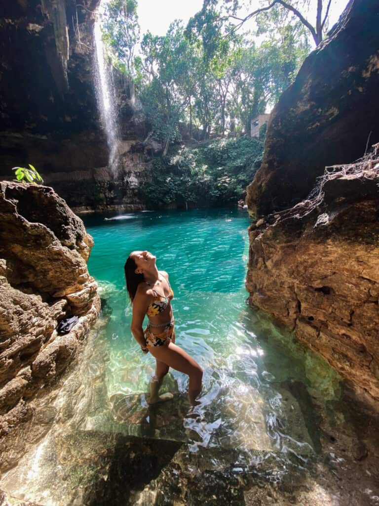 woman in bikini standing in crystal clear blue cenote waters with waterfall