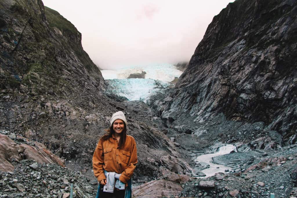 woman in orange flannel and winter hat standing in front of rock and glacier formation in New Zealand