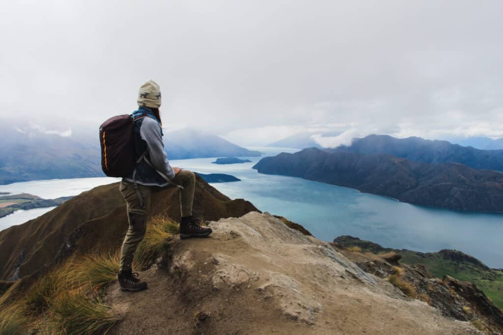 young solo traveler woman standing on top of Roys Peak mountain overlooking lakes in New Zealand on a misty day
