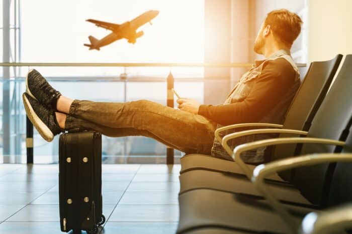 man sitting on airport chair with feet on luggage looking at airplane