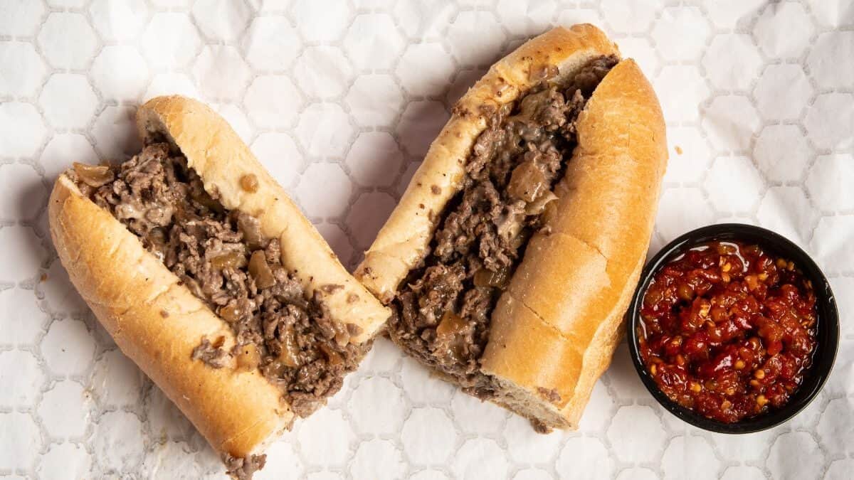 a philly cheesesteak cut in half on white paper with red dipping sauce
