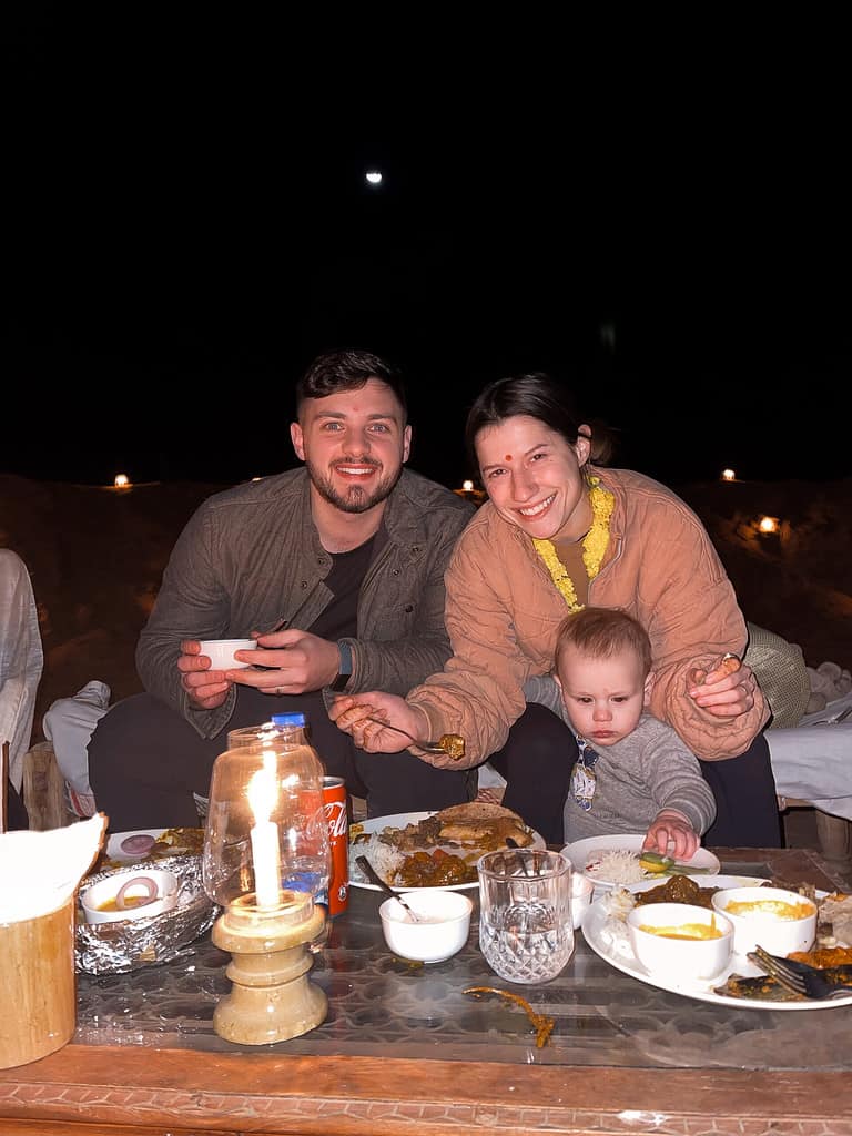 father in dark jacket mother in pink jacket and small toddler eating dinner at night outside