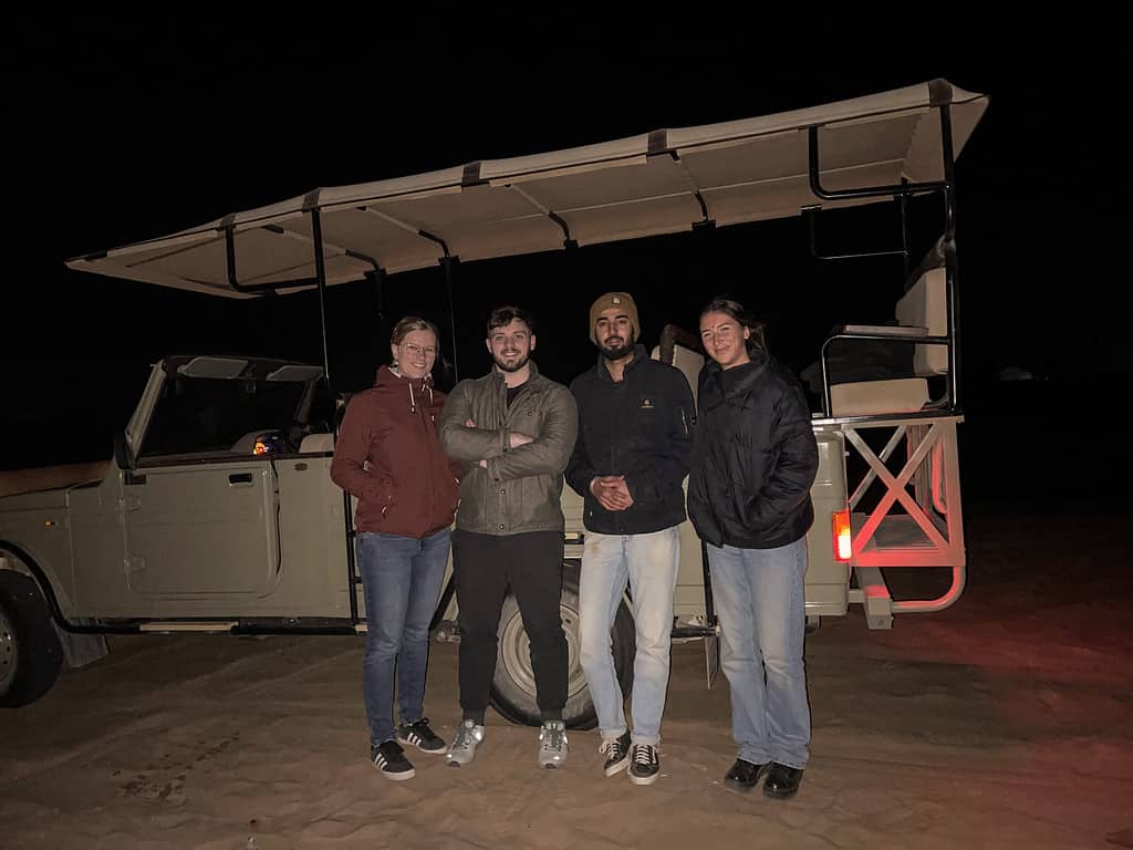 four world travelers standing in front of jeep at night