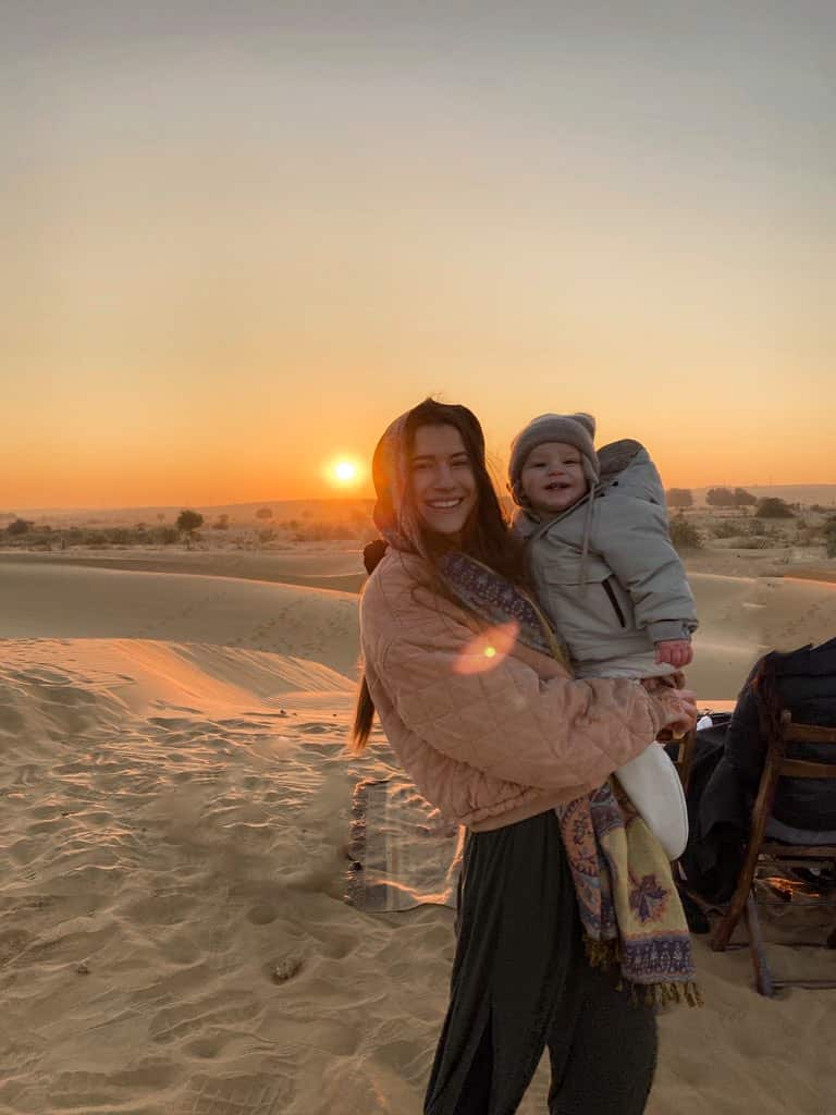 mother bundled in jacekt and scarf holding baby in winter jacket and hat in the desert during sunrise