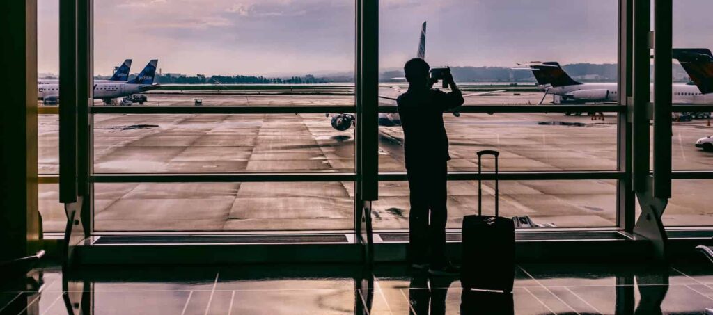 traveler with suitcase standing in front of glass while taking photo of plane