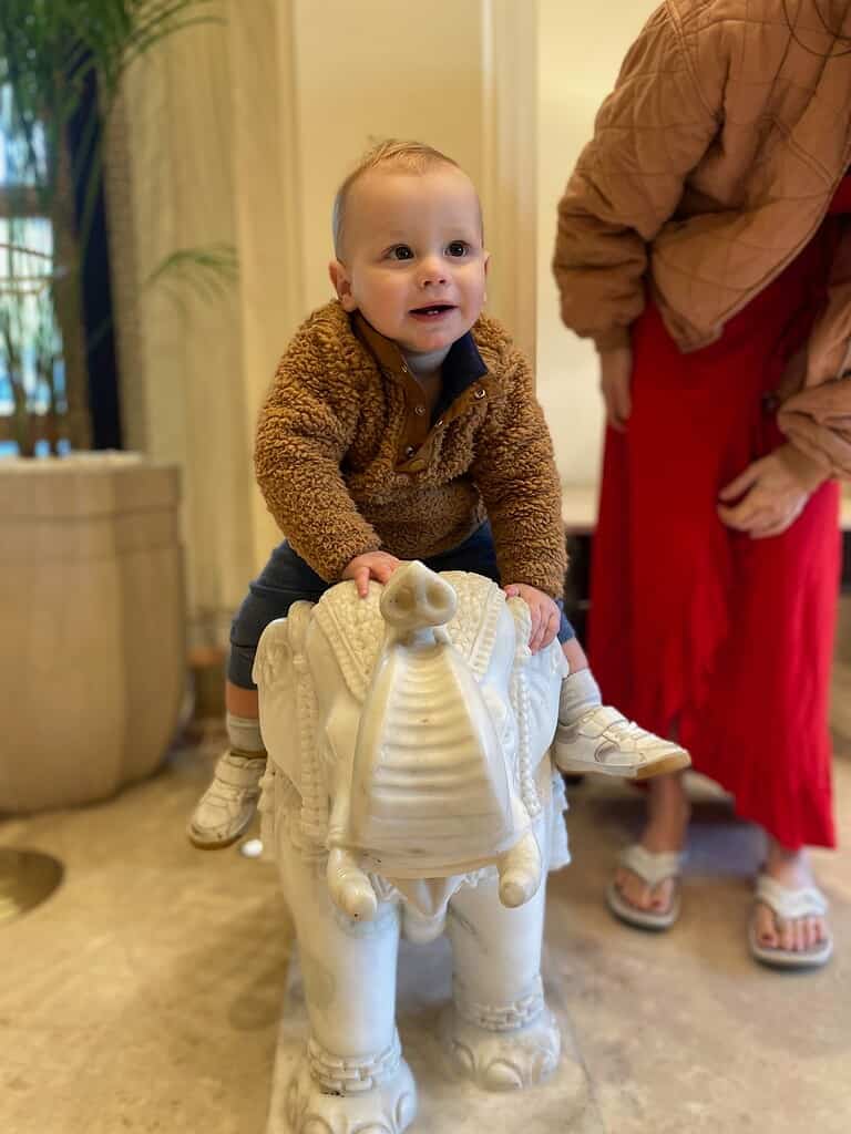 adorable toddler in fuzzy jacket riding a small white marble elephant statue in Indian hotel lobby