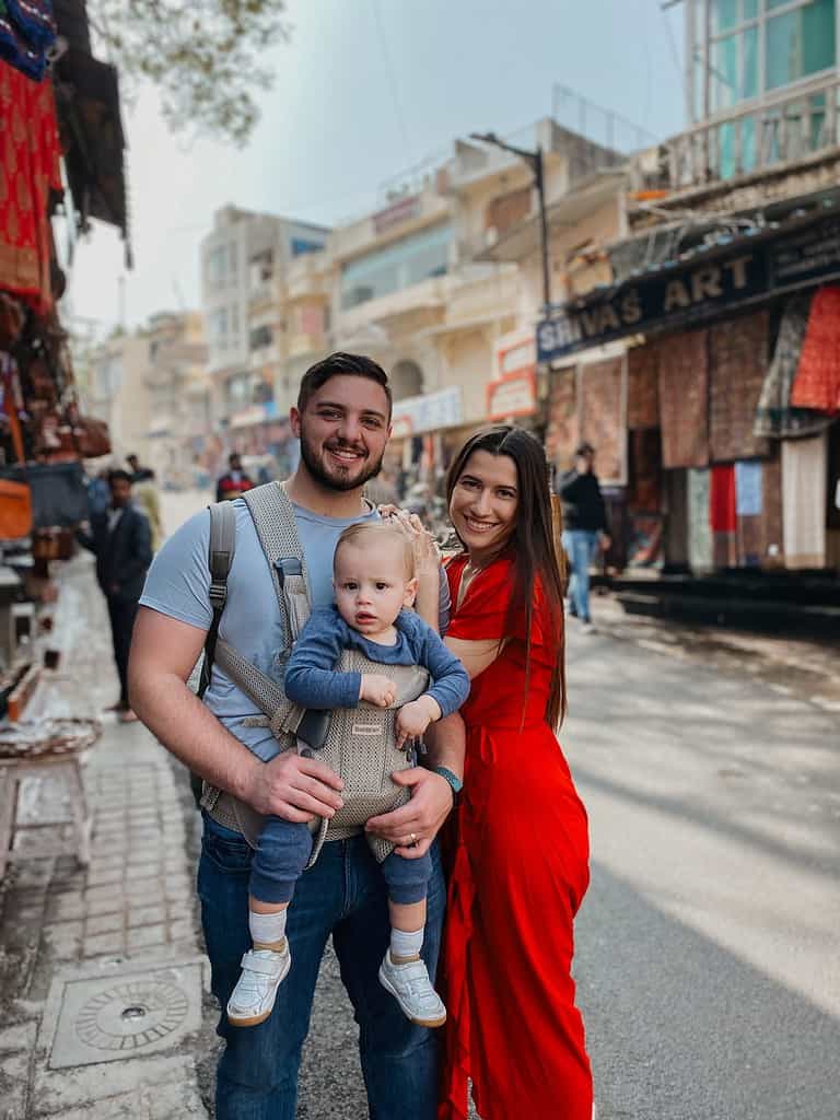 mother in red dress and father with child in a body carrier smiling in the streets of India