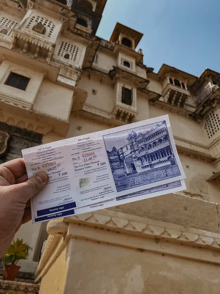 person holding white entrance tickets in front of udaipur city palace wall