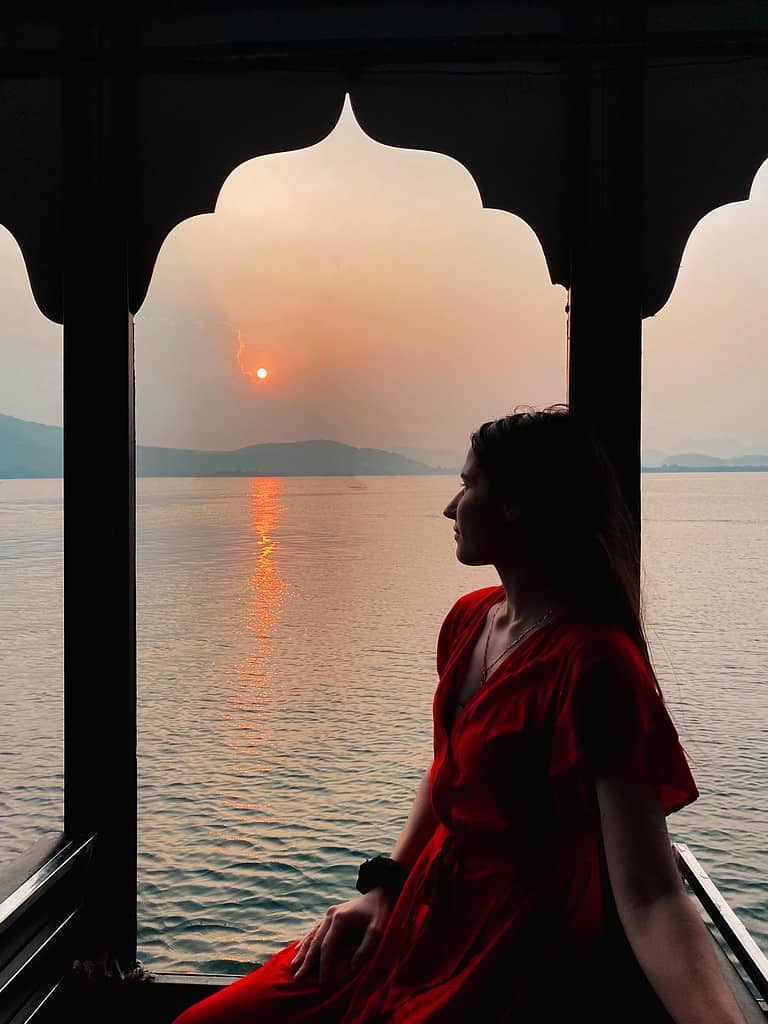 silhouette of woman in red dress sitting under a boat archway overlooking a warm lake sunset