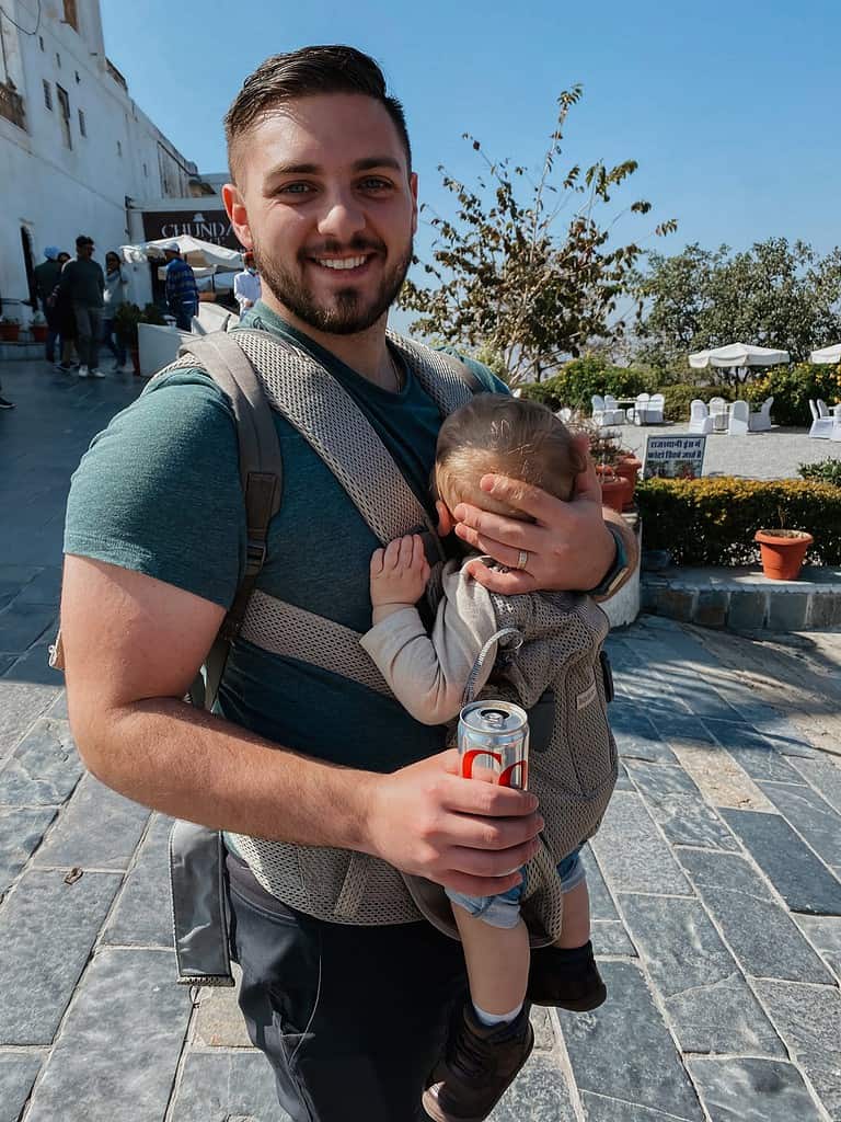 father in green shirt drinking a coca cola with sleeping baby strapped to chest