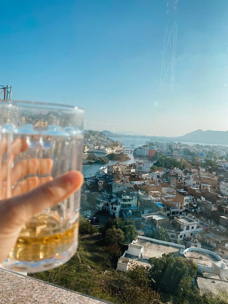 person holding a clear glass of beer on rooftop restaurant overlooking city of Udaipur with clear blue sky