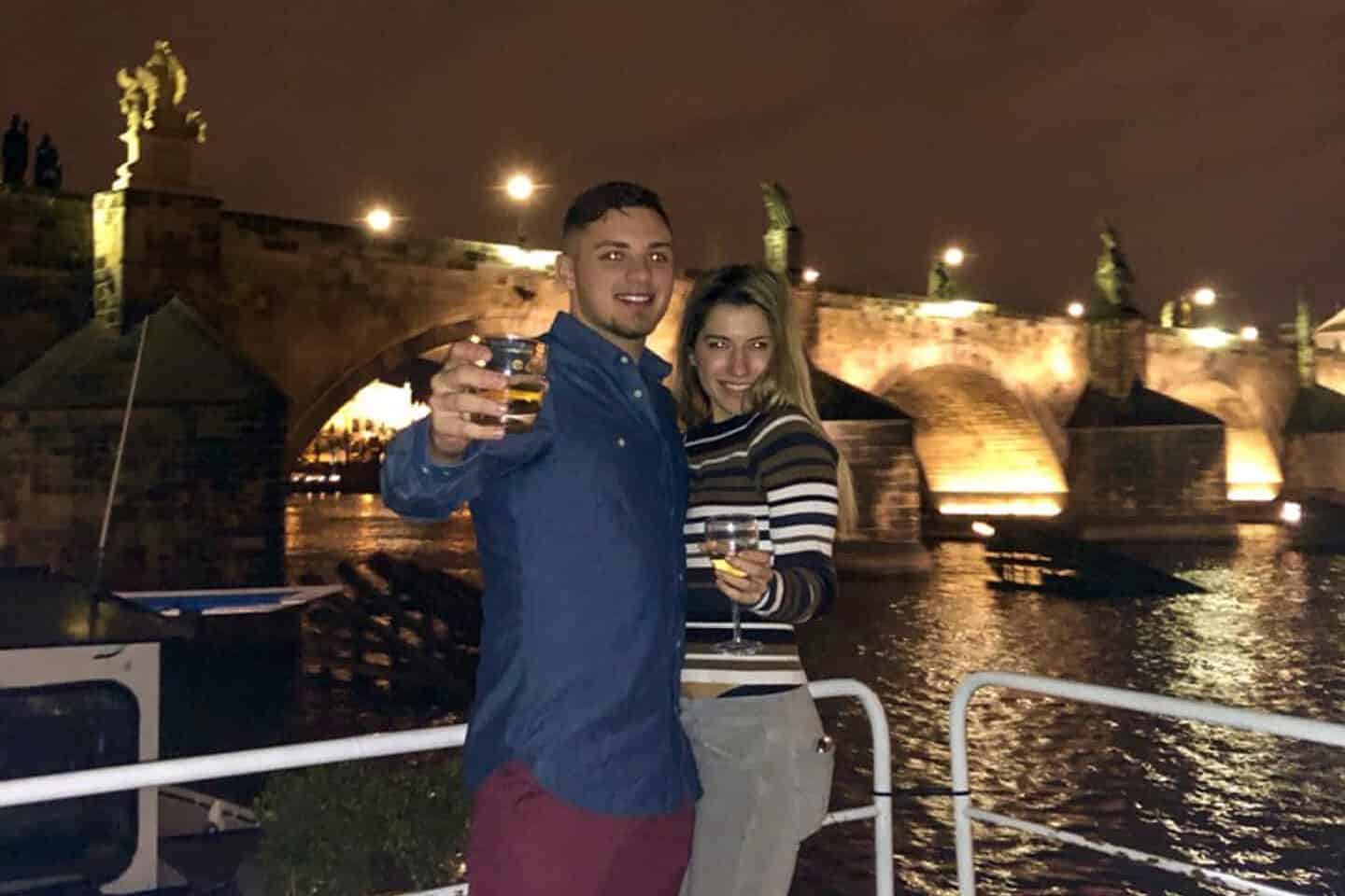 Man and woman raising champagne glasses on a night river cruise with bridge in background