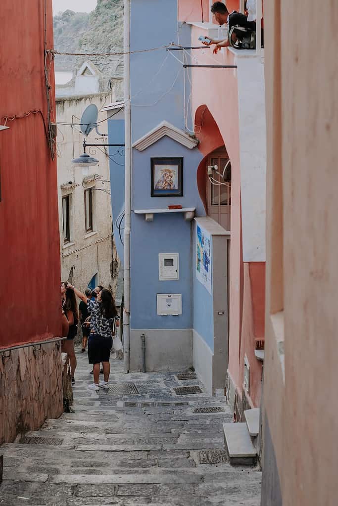 woman in black jacket and black pants standing in colorful italian street of procida during daytime