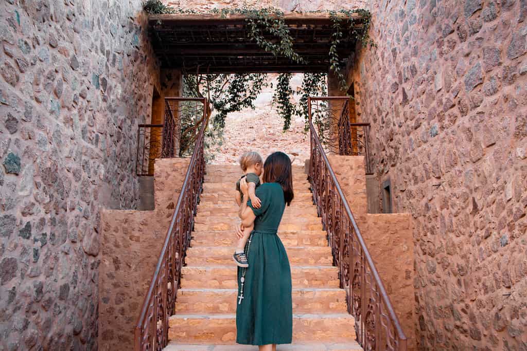 mother and long deep green dress holding toddler son on sand stone stairs with green archway